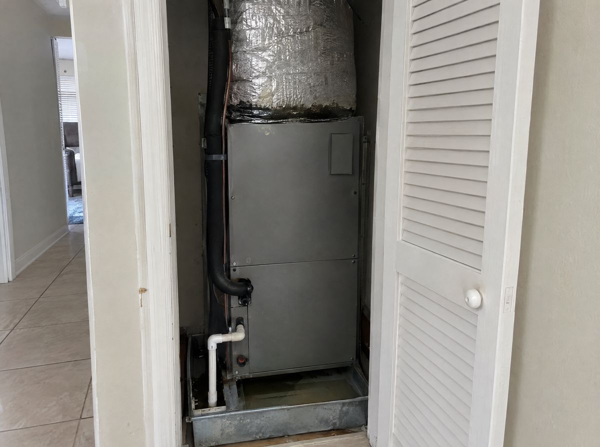 Open hallway closet with an air handler, a white drain line and a little water in the pan underneath.
