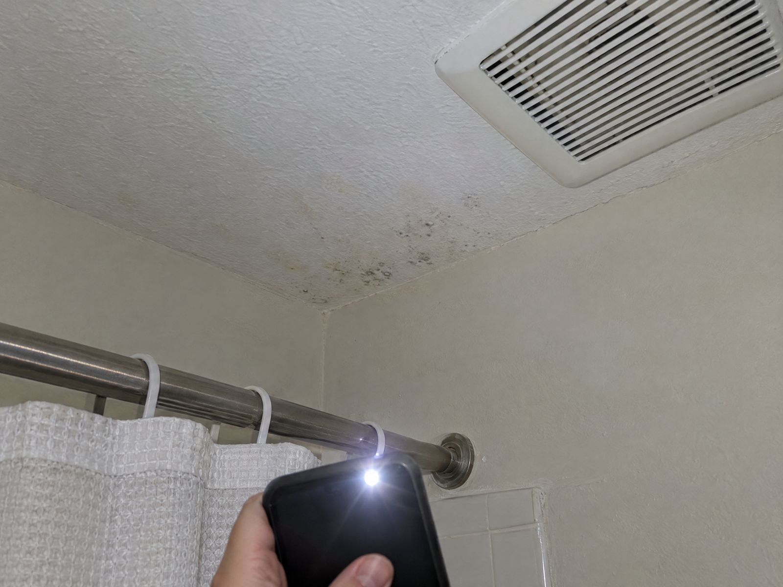 Phone flashlight pointed at small dark spots on a bathroom ceiling near the exhaust fan.