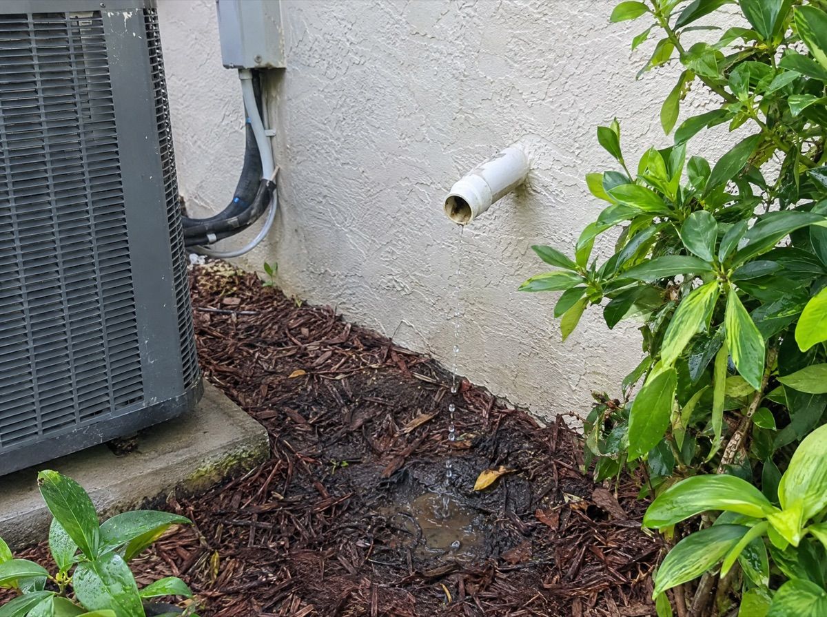 White drain pipe dripping water onto the soil beside an outdoor air conditioning unit.