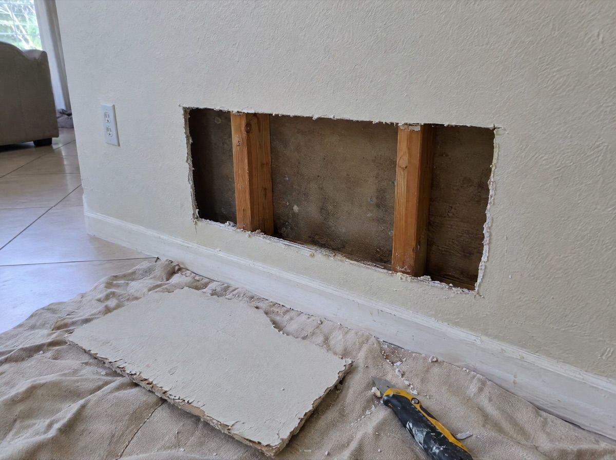 Section of drywall cut out low on a wall, showing the studs and the back of the wall.