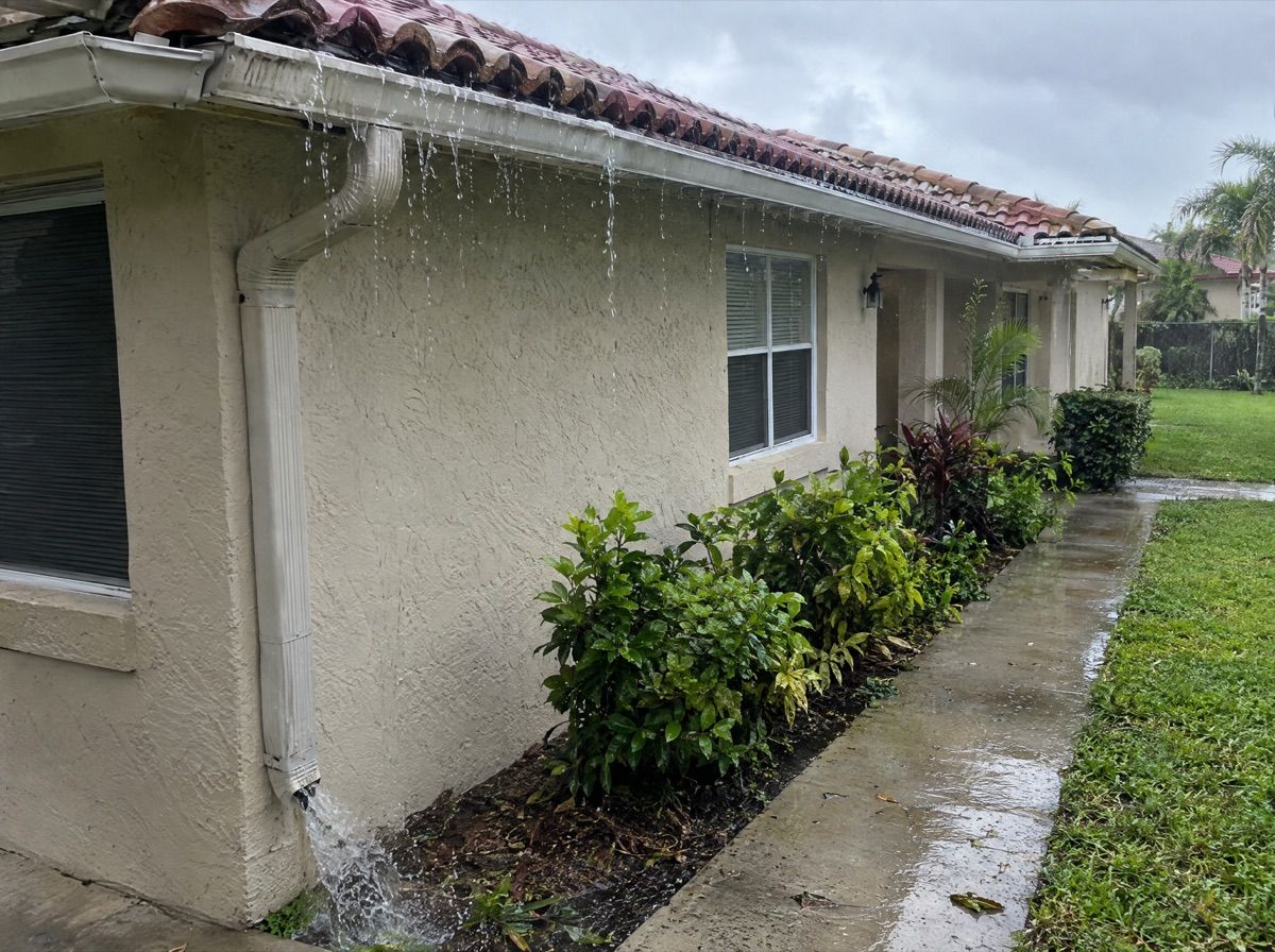 Rainwater overflowing a roof gutter beside a stucco house with a tile roof.