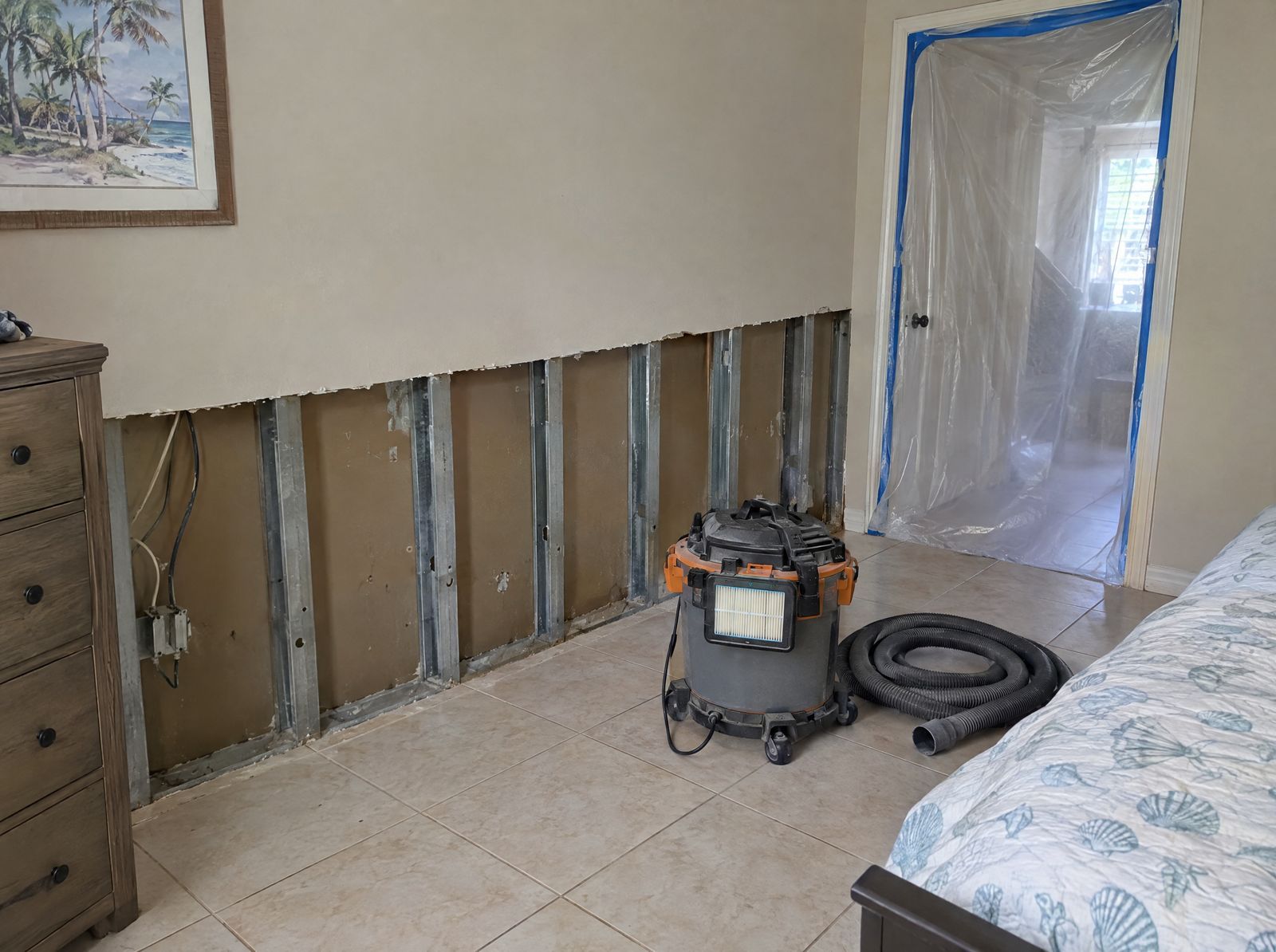 Bedroom wall with the lower drywall cut out, a vacuum on the floor and a doorway covered with plastic sheeting.