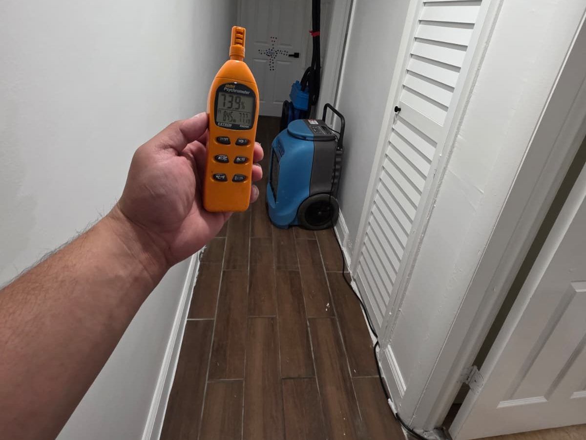 Hand holding a humidity meter in a hallway with a dehumidifier in the background.
