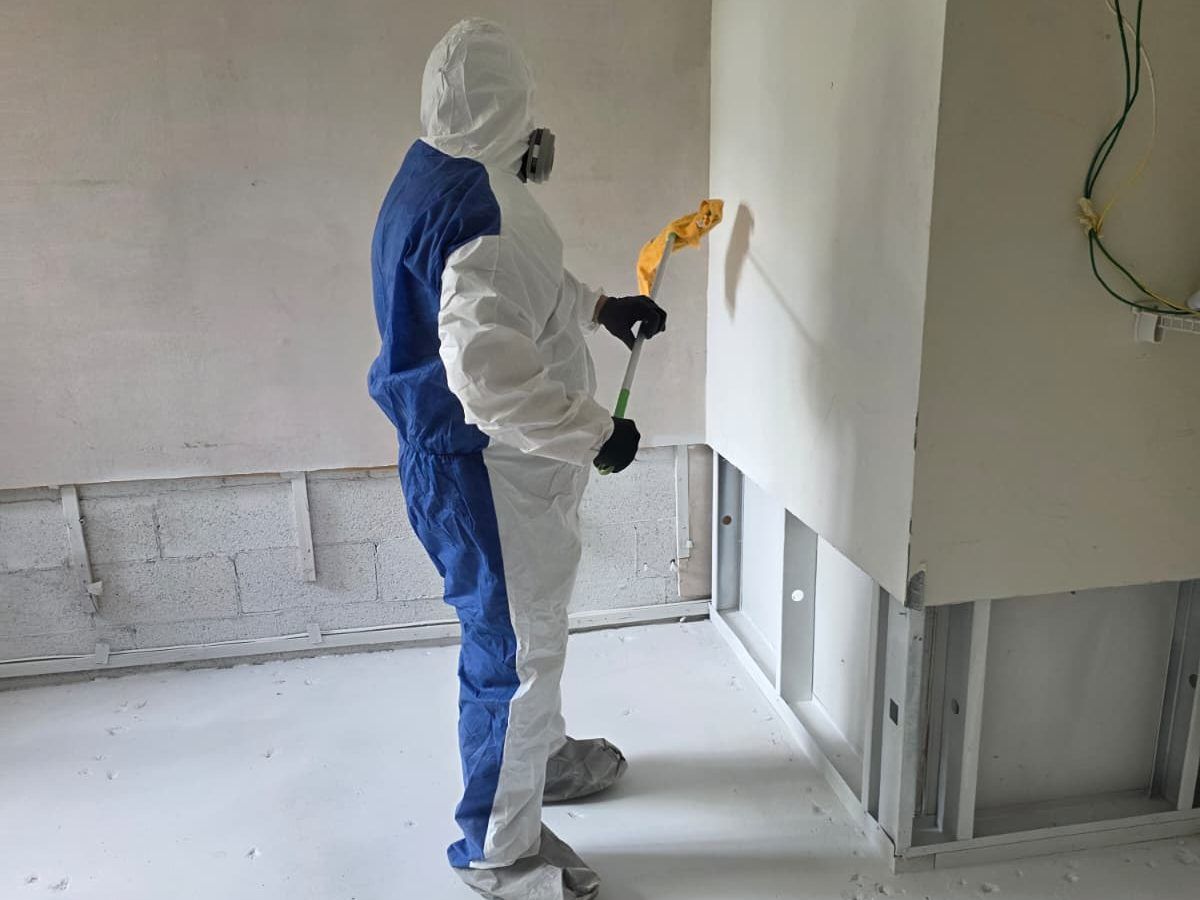 Worker in a protective suit cleaning a wall in an empty room with the lower drywall removed.