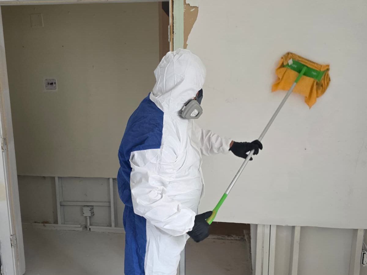 Worker in a protective suit and respirator wiping a wall with a pad on a pole.