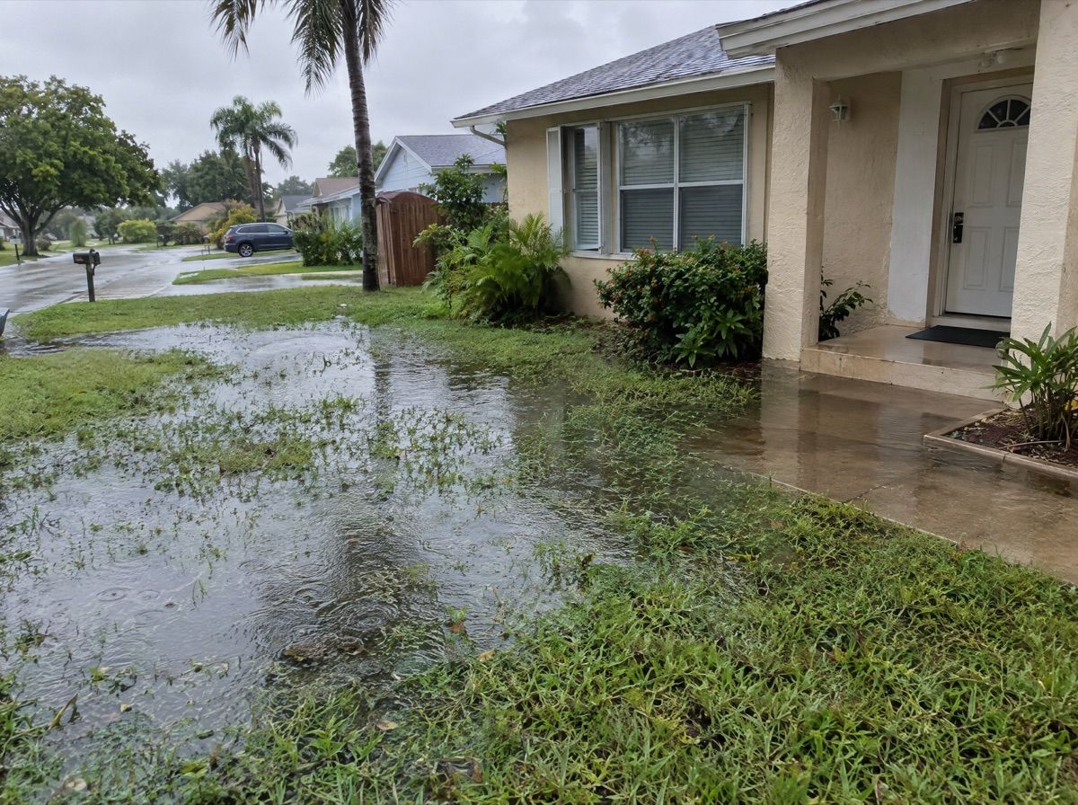 Shallow street floodwater reaching the front walkway of a stucco house after a storm.