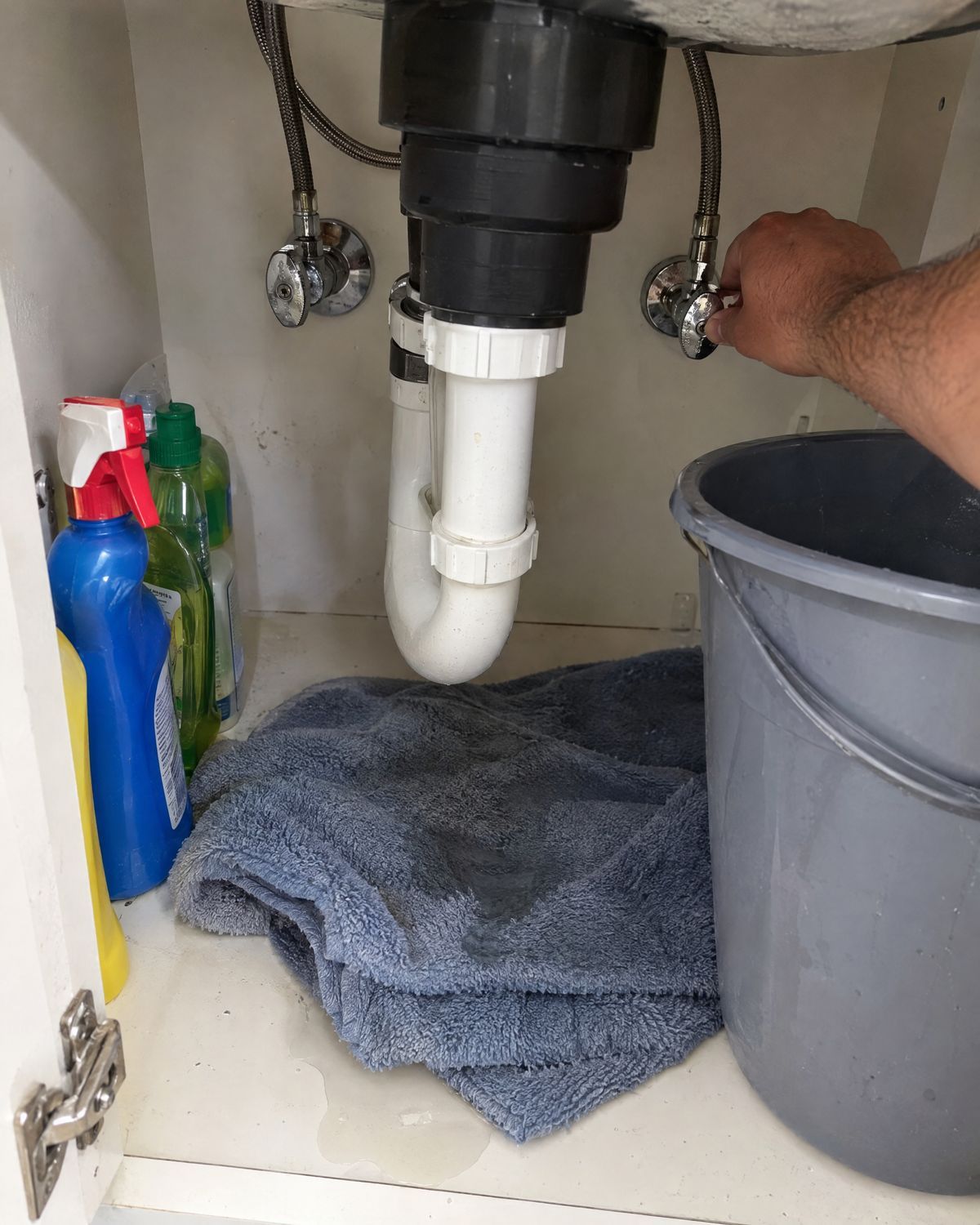 Hand closing the water shut-off valve under a kitchen sink, with a towel and a bucket catching a leak.