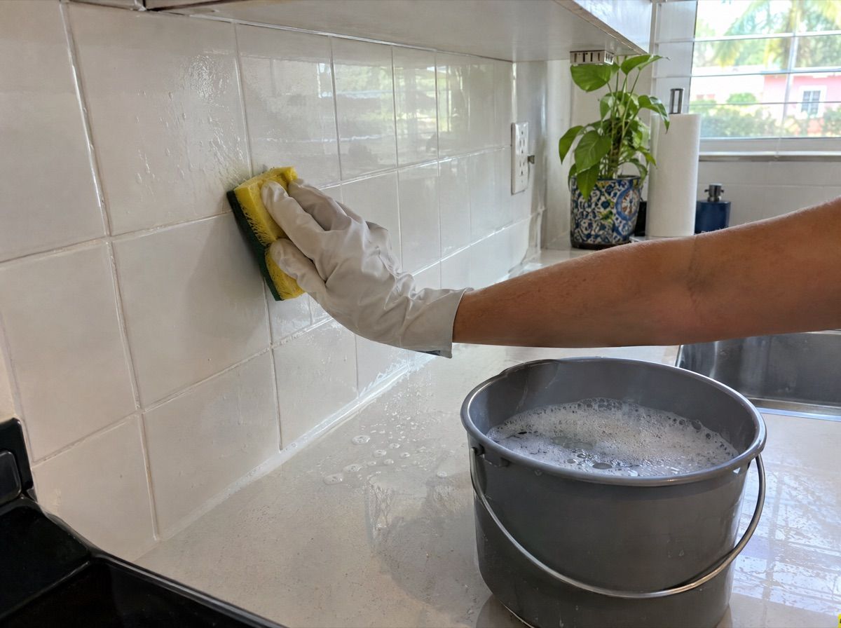 Gloved hand wiping a tiled kitchen counter with a sponge, next to a bucket of soapy water.